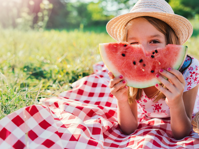A girl in a straw hat smiles while holding a slice of watermelon on a picnic blanket.