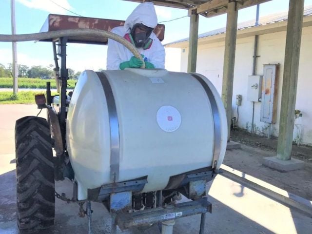 A person in protective gear operates a tractor with a large spray tank.