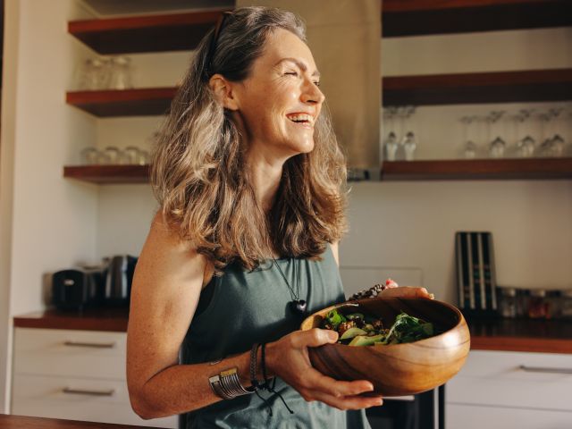 Smiling woman in a kitchen holding a wooden bowl of salad. Natural light fills the space.