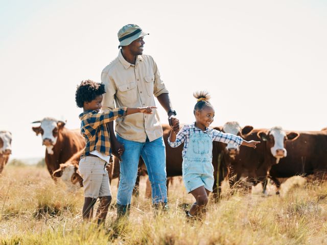A man and two children joyfully point while walking in a field with cows in the background.