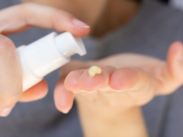 A hand dispensing a small amount of cream from a white pump bottle onto an open palm.