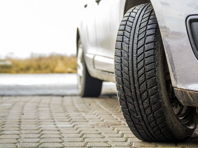 Close-up of a textured car tire resting on a paved surface, with a blurred vehicle in the background.