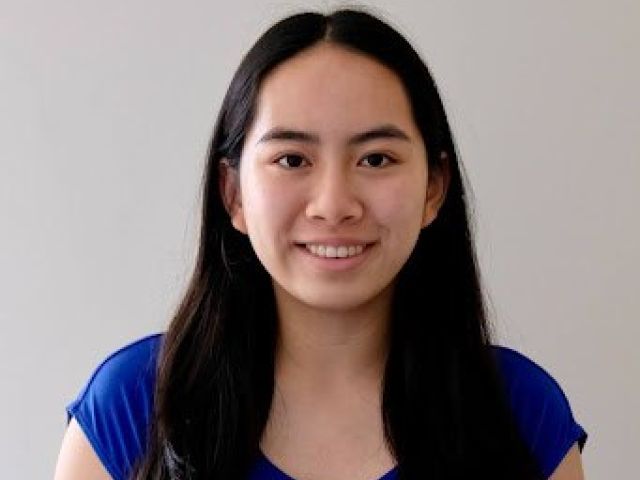 Woman with long black hair smiling, wearing a blue shirt against a neutral background.