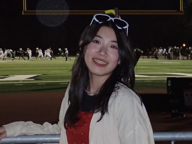 Young woman smiling, wearing sunglasses, at a nighttime sports event with a field in the background.