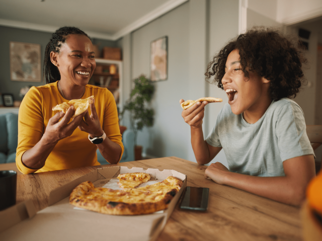 A woman and a boy enjoy pizza together at a table, laughing and sharing a joyful moment