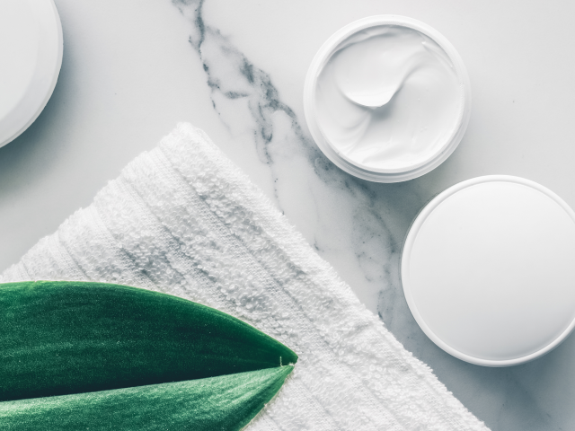 Two white cream jars on a marble surface next to a green leaf and a towel.