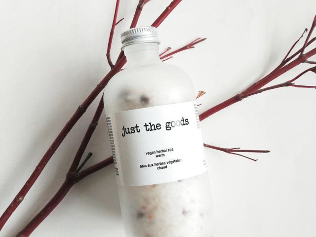 Transparent bottle labeled "just the goods" with herbal bath soak, surrounded by twig branches.
