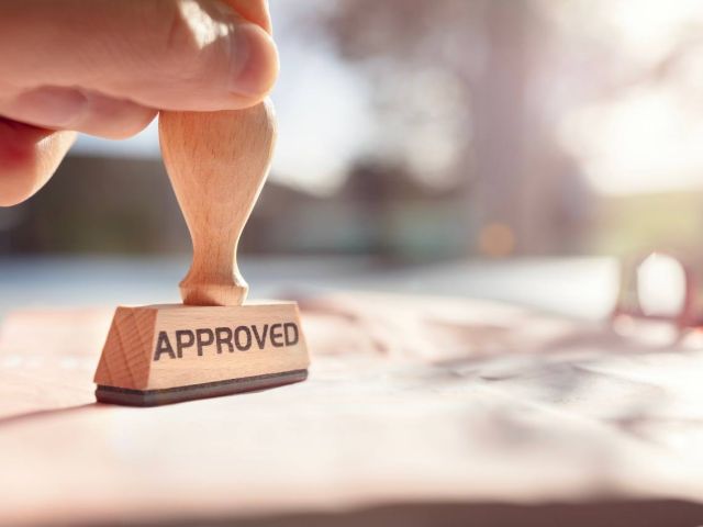 A hand holding a wooden stamp marked "APPROVED" above a document on a sunny surface.