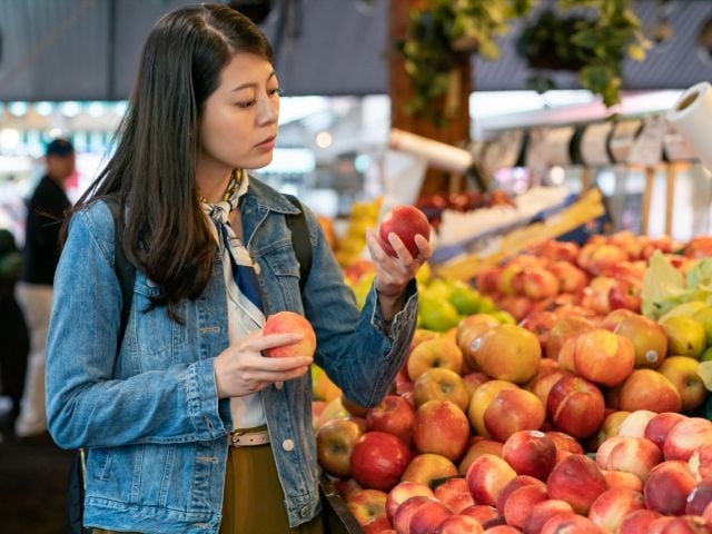 Photo: woman in denim jacket examining a red apple at a busy market stall