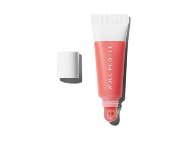 Photograph of open coral lip gloss tube labeled WELL PEOPLE, white cap beside on white background