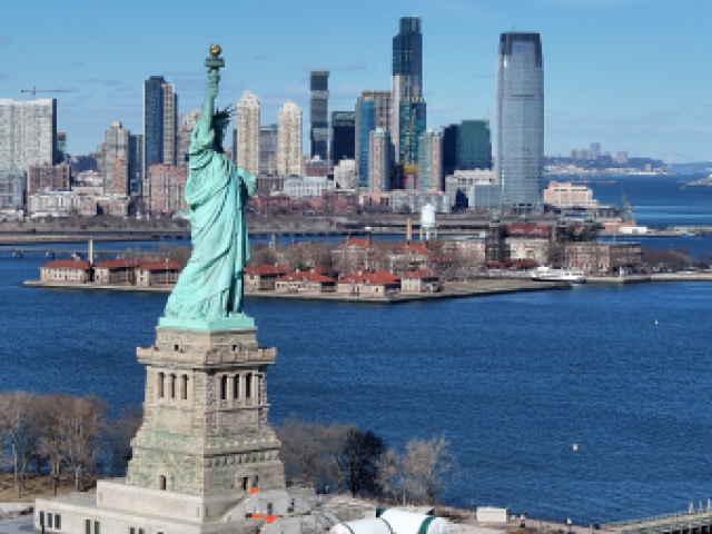 Photograph of Statue of Liberty with Ellis Island and Manhattan skyline across blue harbor