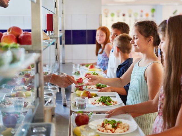 Photograph of smiling children in a school cafeteria receiving trays with salads and apples.