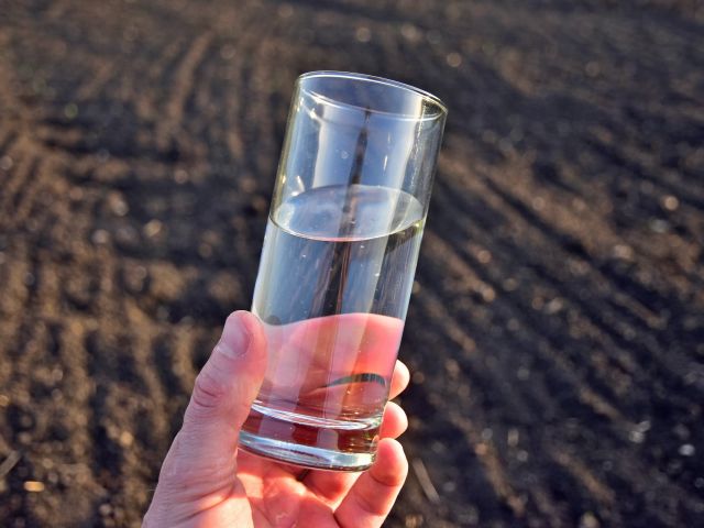 Photograph of hand holding clear glass of water above plowed field