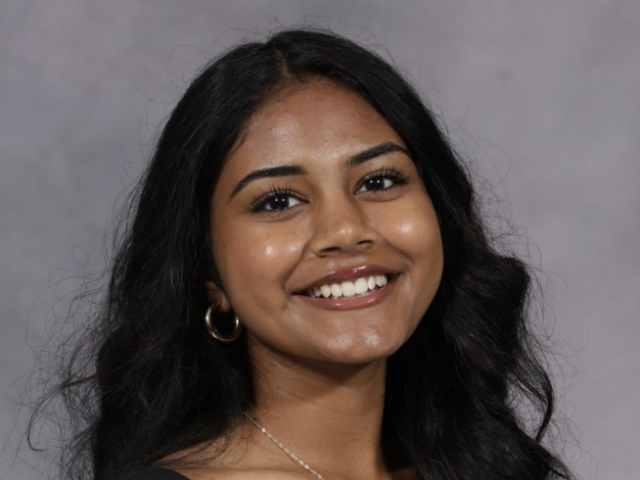 Photograph of a smiling young woman with long dark hair, gold hoop earring, gray backdrop