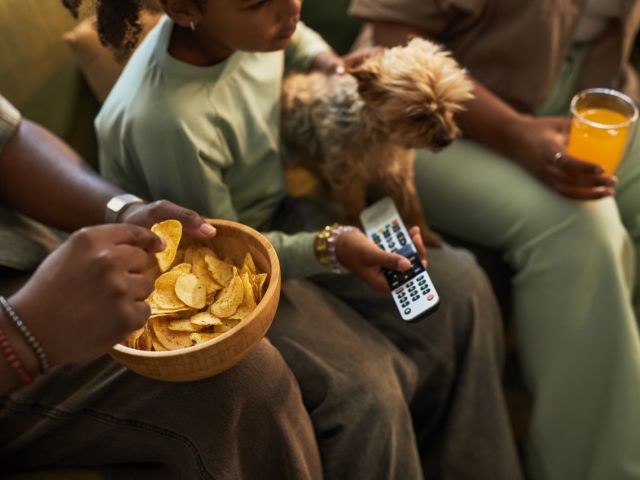 Photo: family sharing chips on couch with small dog, TV remote and glass of orange juice