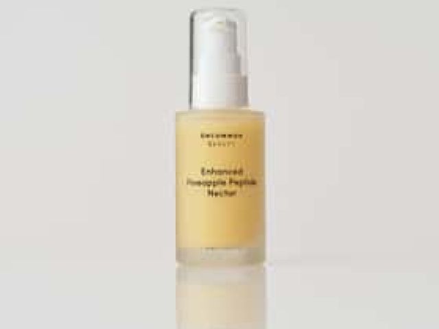 Photograph: clear pump bottle with pale yellow serum on white surface, subtle reflection.
