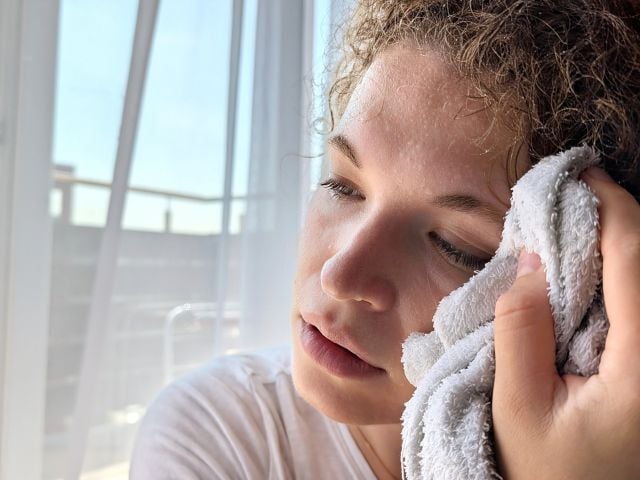 Photograph of a young woman with eyes closed pressing a towel to her cheek by sunlit curtains.