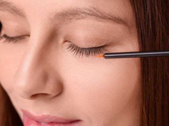 Close-up photograph of woman with closed eye applying liquid eyeliner to upper lashes