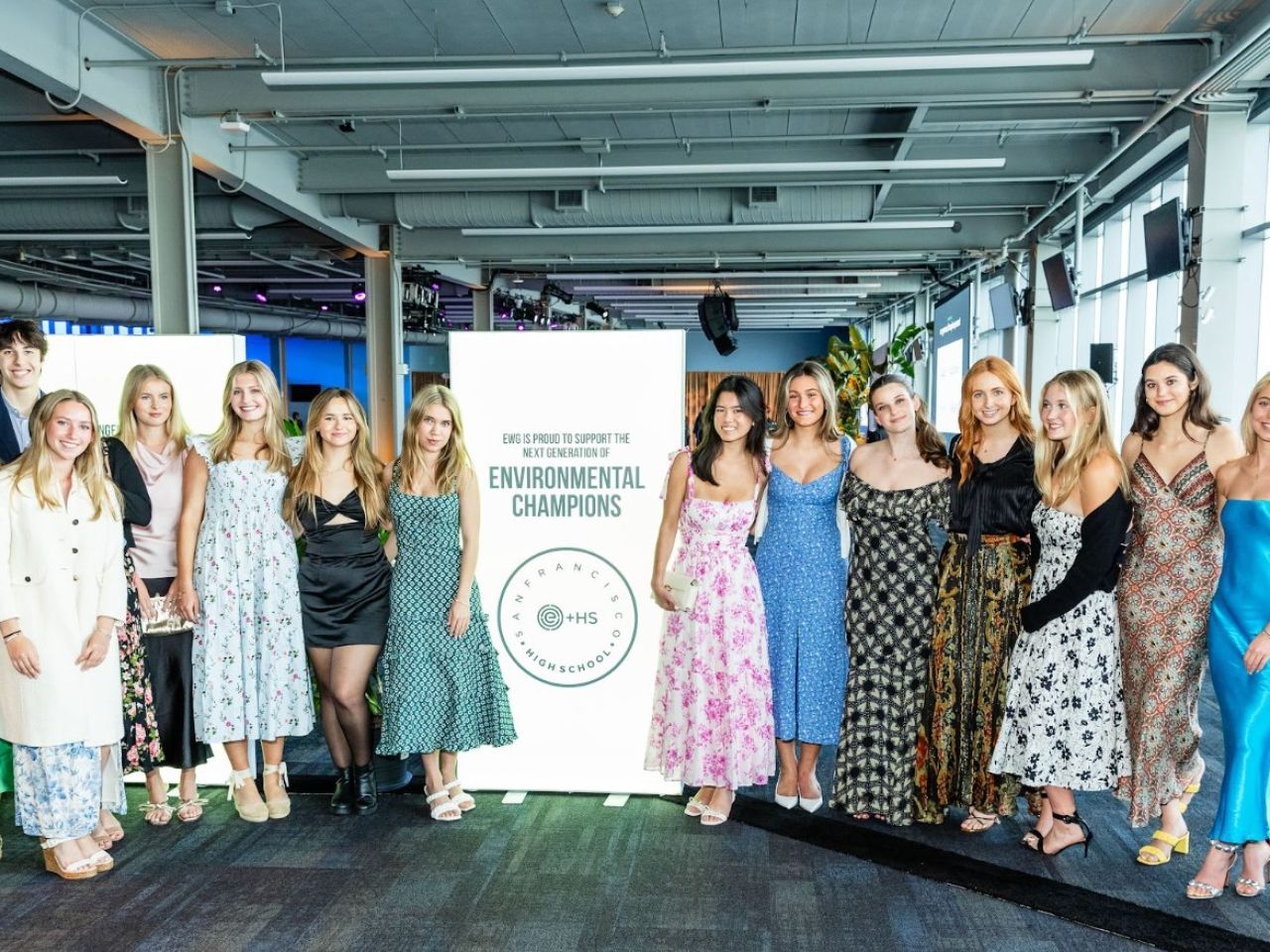 A group of young women in various dresses poses in a modern event space with large windows.