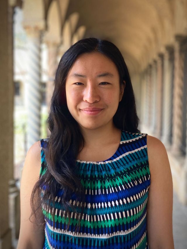 A young woman with long dark hair smiles, wearing a colorful patterned top, in a corridor.
