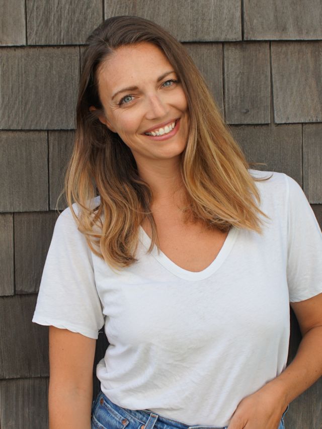 Woman with long, wavy hair and a bright smile, standing against a wooden wall.