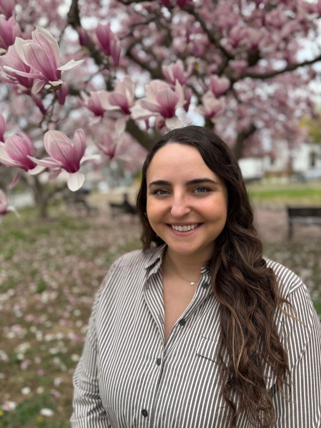 Young woman with long hair smiles amid blooming magnolia trees in a park.