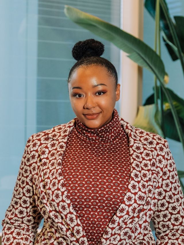 A woman with a bun hairstyle wears a patterned blazer over a maroon turtleneck, smiling.