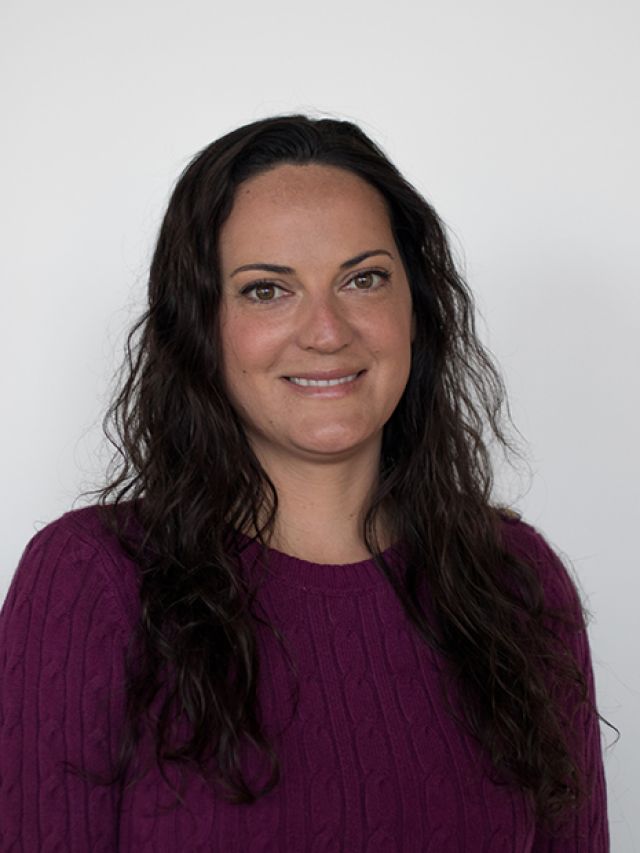 Smiling woman with long, dark, wavy hair wearing a purple sweater against a white background.