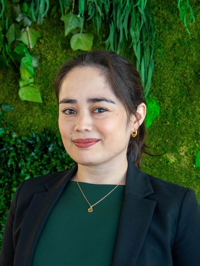 A woman in a black blazer smiles against a green, plant-filled backdrop.