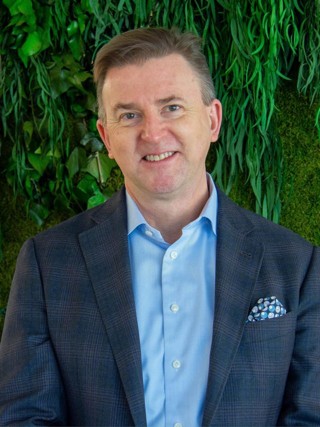 A smiling man in a blazer stands against a backdrop of green foliage.