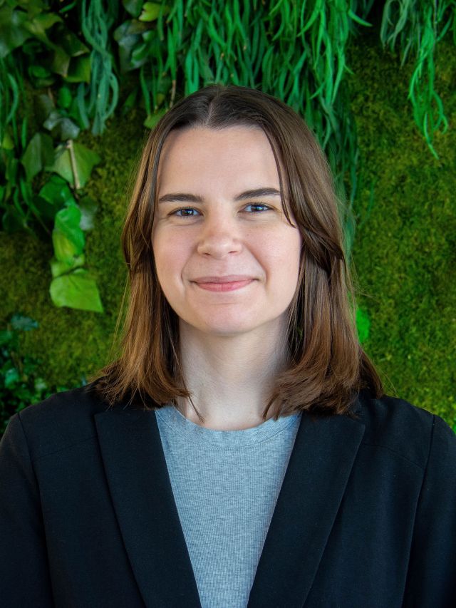 A young woman with shoulder-length brown hair smiles, dressed in a black blazer against a green wall.