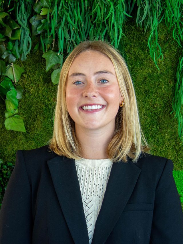 Smiling woman with blonde hair in a black blazer, posing against a green plant background.