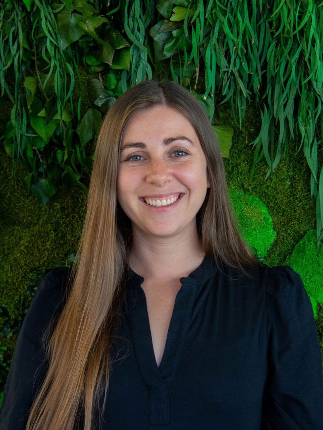 Young woman with long brown hair smiling in front of a green plant backdrop.
