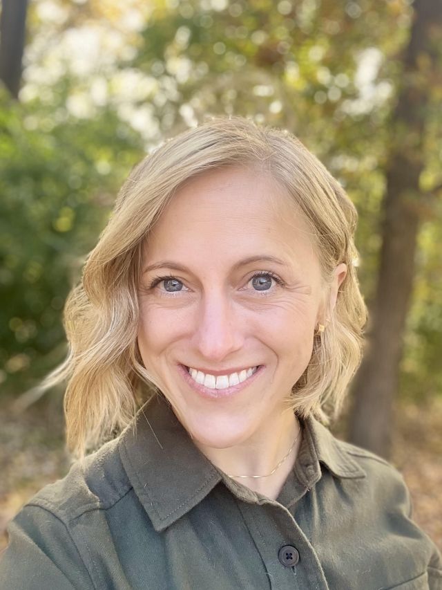 Smiling woman with wavy blonde hair standing outdoors among green trees.