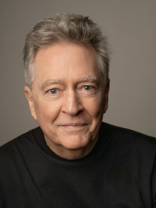 Headshot photograph of man with gray hair, black sweater, neutral background