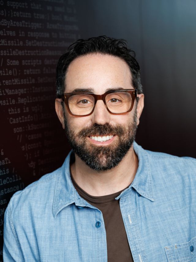 Photograph: smiling bearded man with glasses in blue shirt, dark background with code text