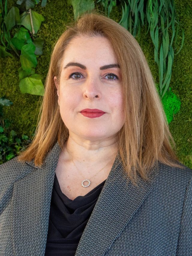 Photograph: woman with shoulder-length light brown hair, gray blazer, against green plant wall