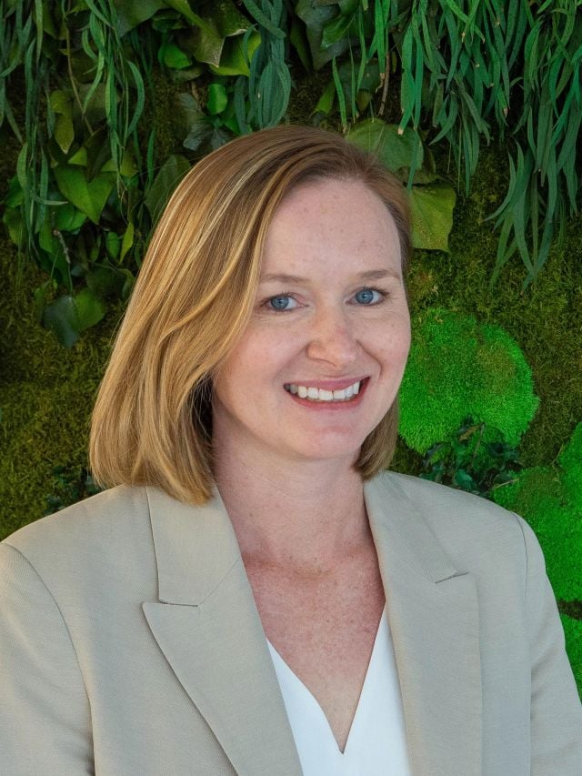 Photograph of smiling woman with blonde bob in beige blazer against green living wall