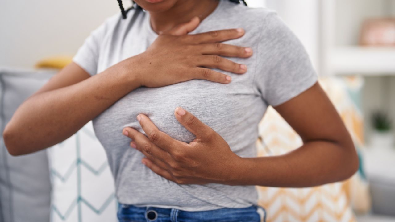 A woman sitting on a couch holds her chest, expressing discomfort or concern.
