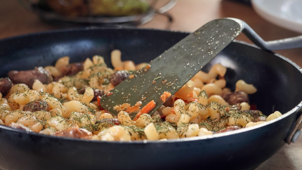 A skillet filled with pasta, vegetables, and herbs being stirred with a spatula.