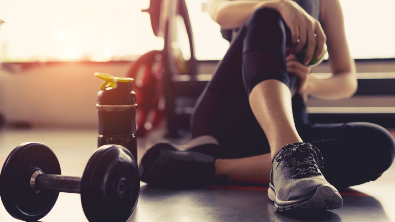 A person sits on a gym floor beside a dumbbell and a water bottle, resting.