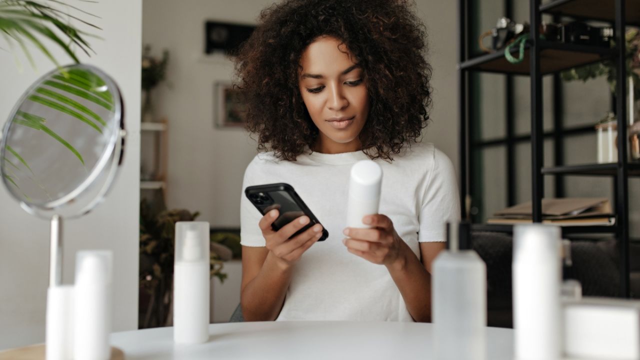 Woman with curly hair checking her phone while holding a skincare product.