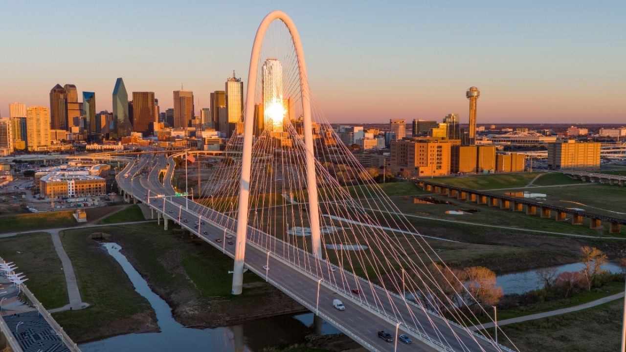 Dallas skyline at sunset with a modern suspension bridge in the foreground.