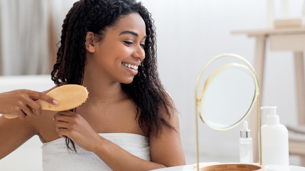 A young woman brushes her curly hair while smiling in front of a mirror.