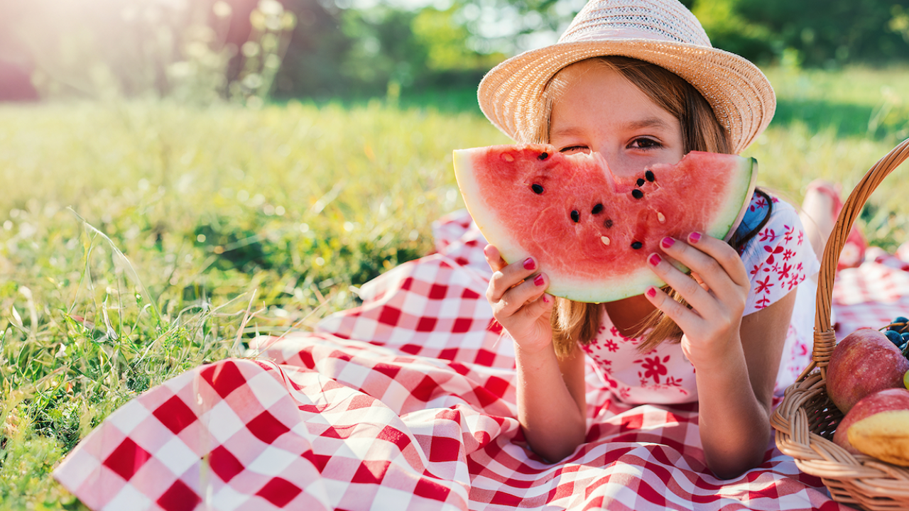 A girl in a straw hat smiles while holding a slice of watermelon on a picnic blanket.