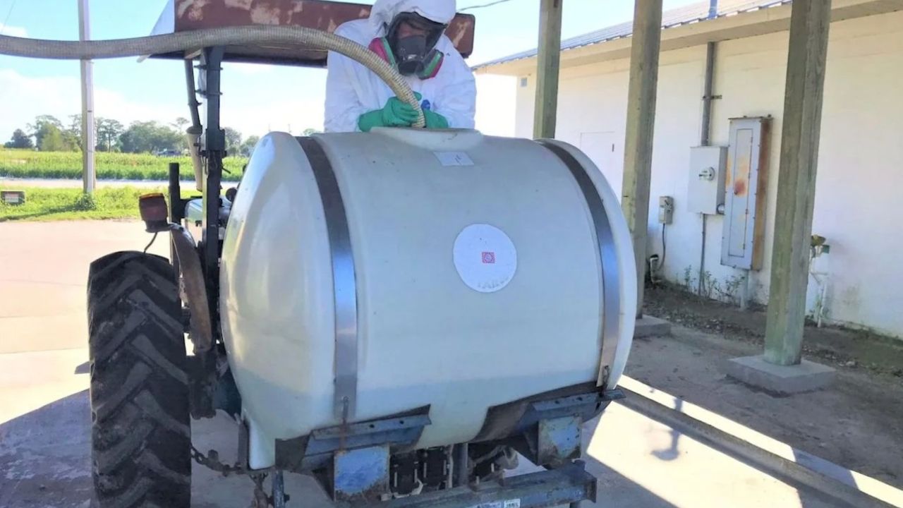 A person in protective gear operates a tractor with a large spray tank.