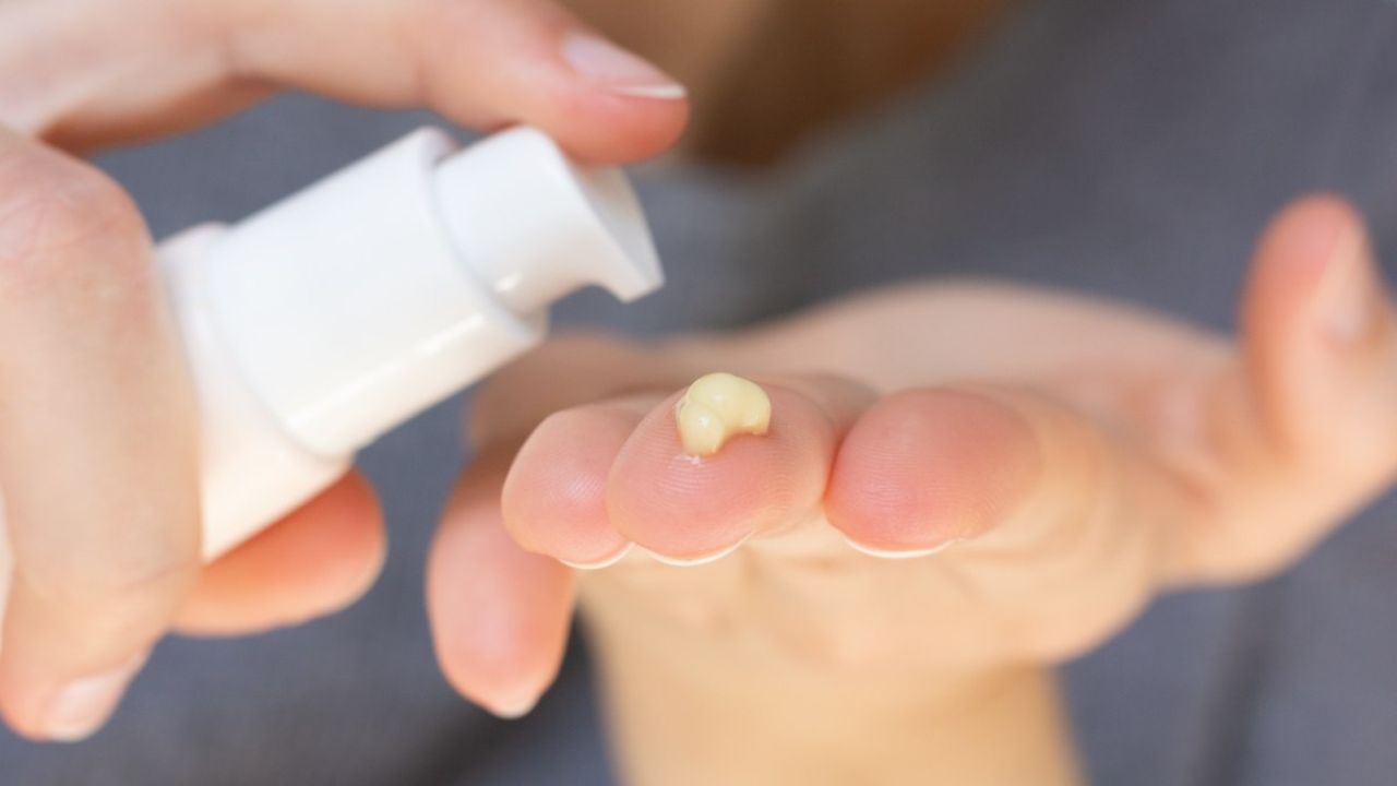 A hand dispensing a small amount of cream from a white pump bottle onto an open palm.