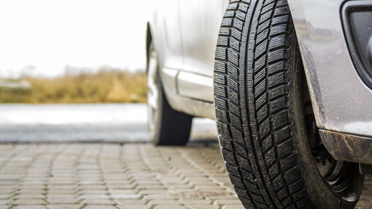 Close-up of a textured car tire resting on a paved surface, with a blurred vehicle in the background.