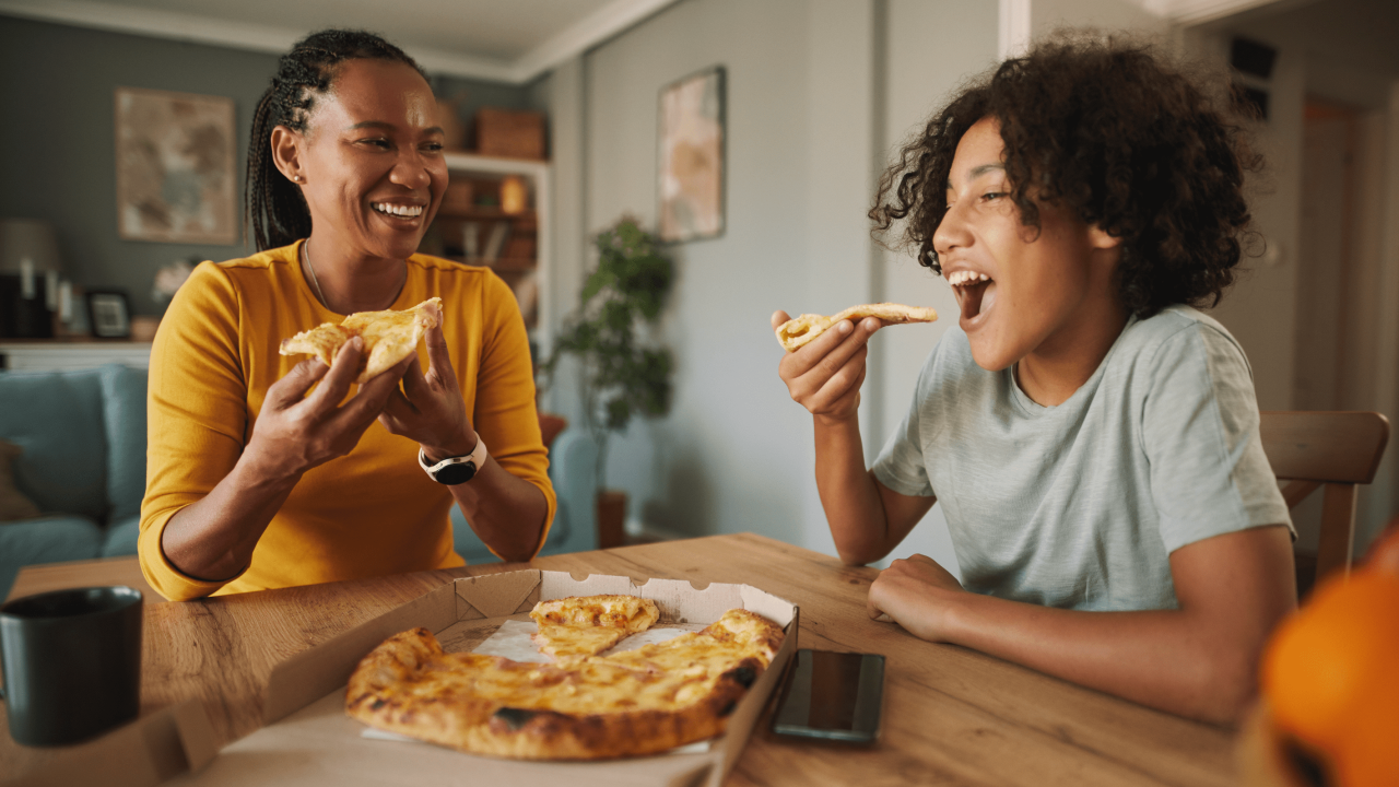 A woman and a boy enjoy pizza together at a table, laughing and sharing a joyful moment