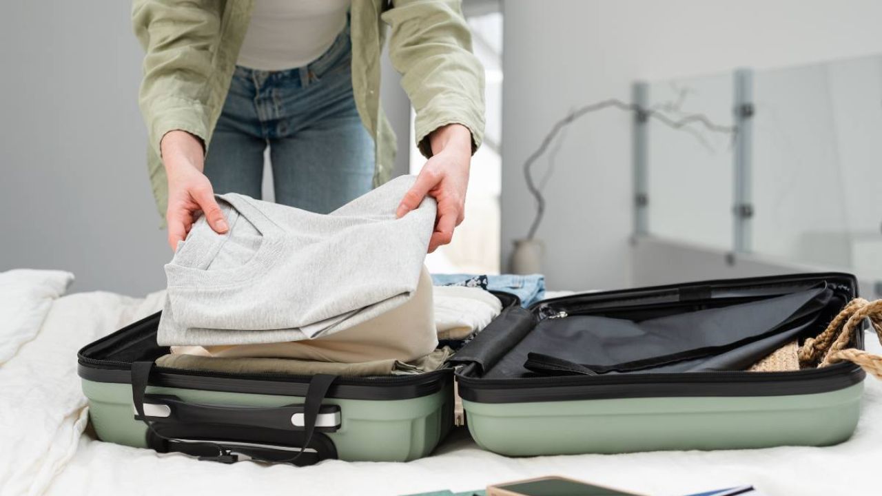 A person packing gray pants into a green suitcase on a bed. A notebook and phone are nearby.
